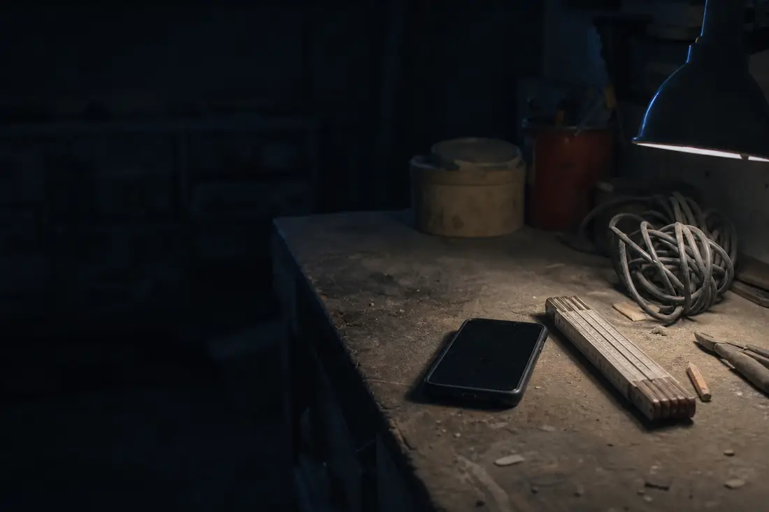 Telefon egy poros munkapadon, mellette colstok és kábel, egyetlen műhelylámpa fényében